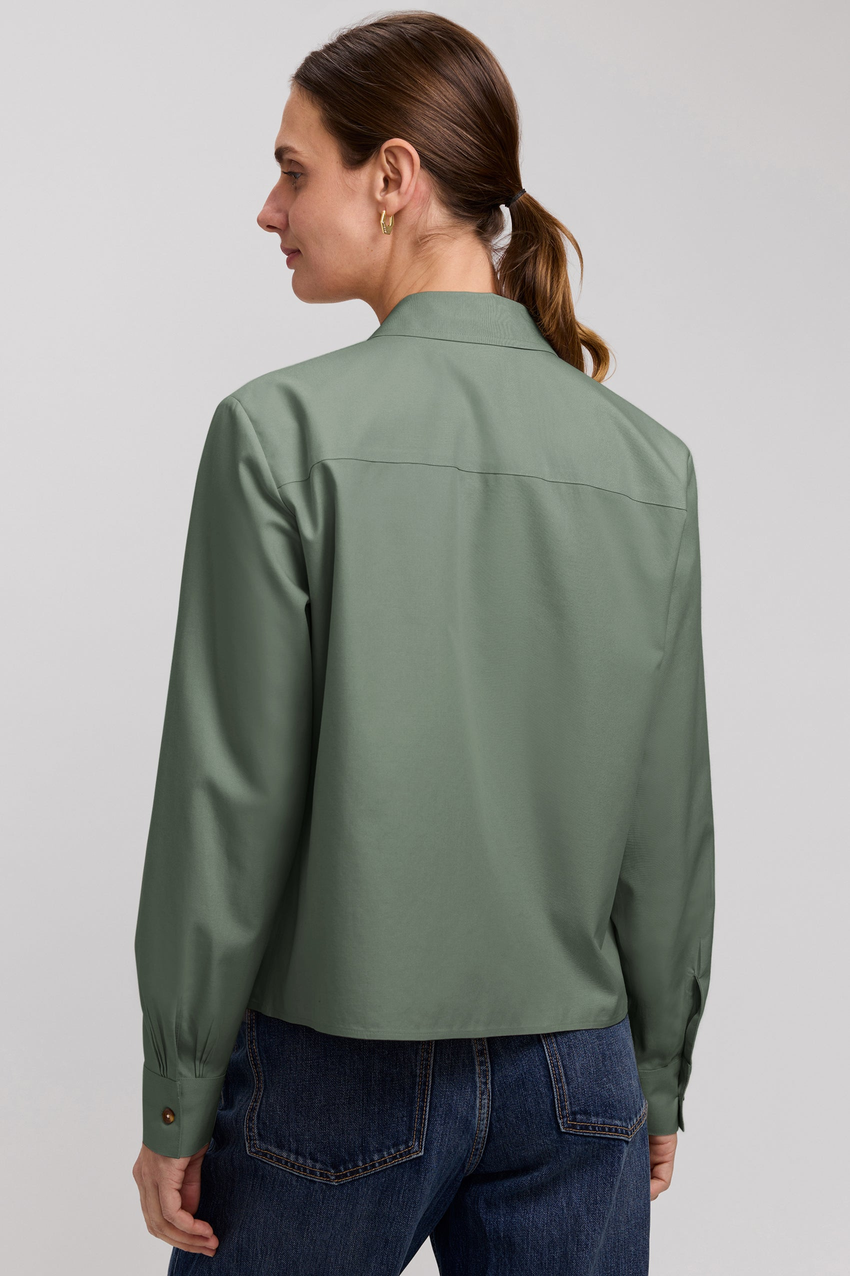 Back view of a woman wearing a sage green long sleeve short shirt with dark blue jeans.