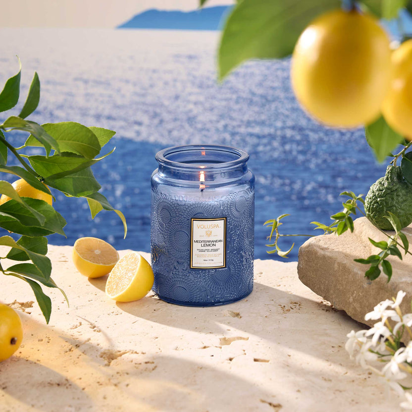 Mediterranean Lemon 18oz Large Jar Candle in blue glass on a stone ledge with lemons and sea view.