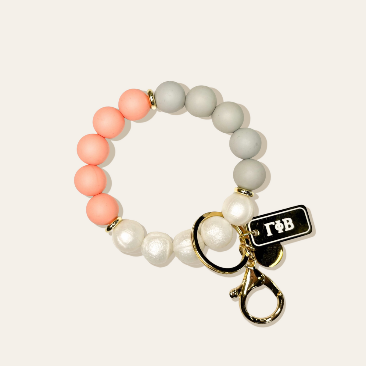 Beaded keychain wristlet with coral, grey, and pearl beads, gold hardware, and a black tag with Greek letters.