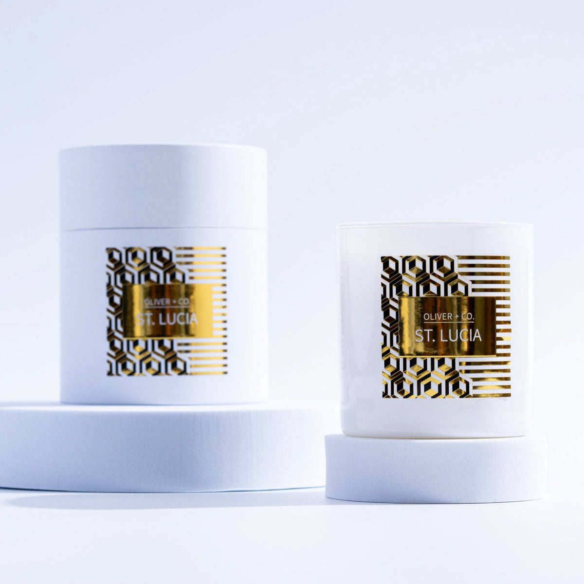 White 12 oz St. Lucia signature candle and its matching cylindrical box with gold geometric labels.