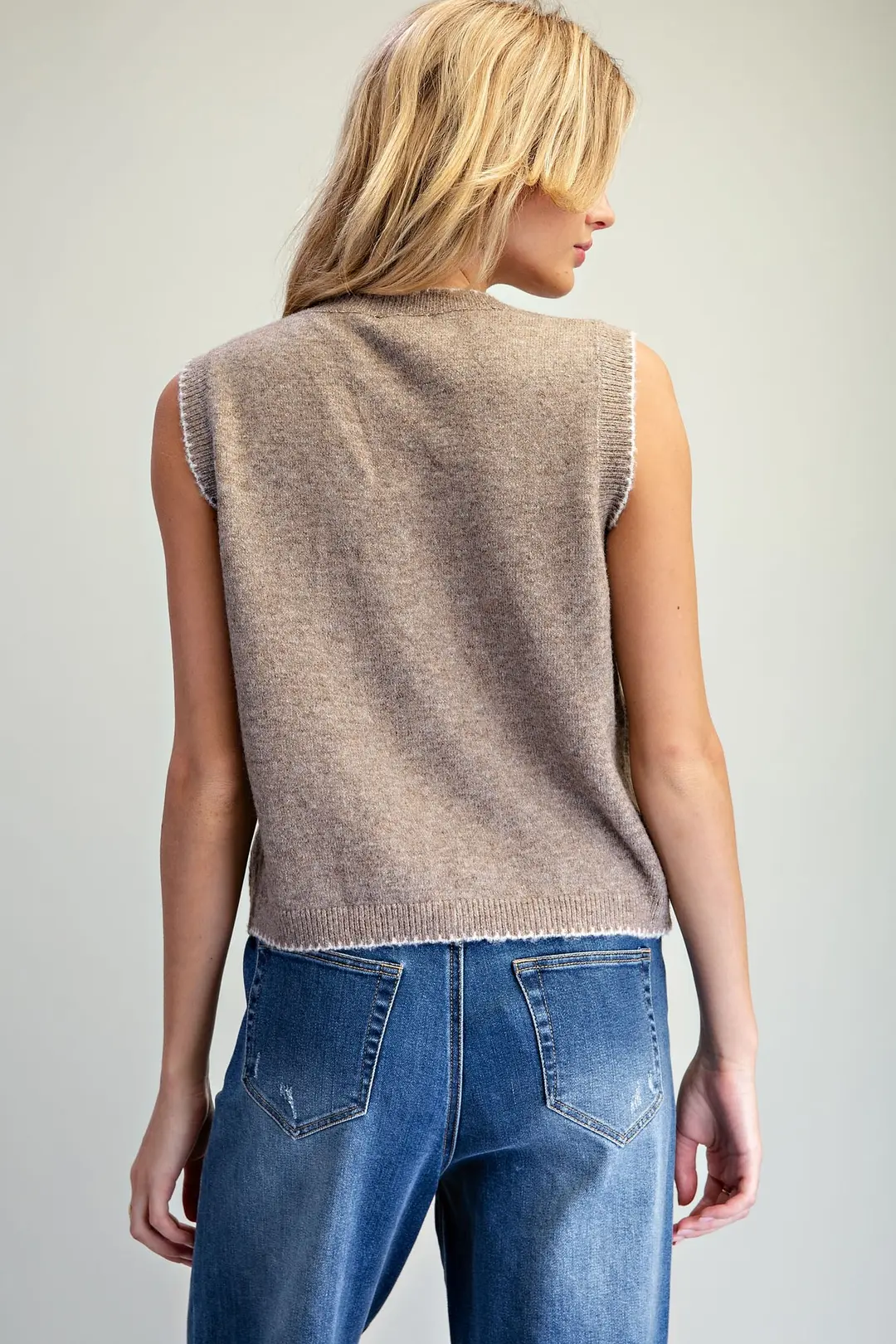 Back view of a woman wearing a brown knit vest with white whipstitch trim and blue jeans.