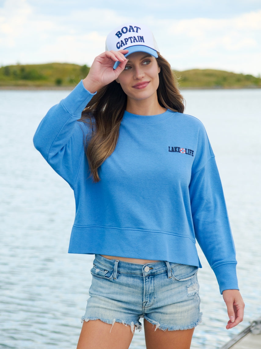 Woman wearing a white and blue Boat Captain trucker hat with a blue sweatshirt by the water.