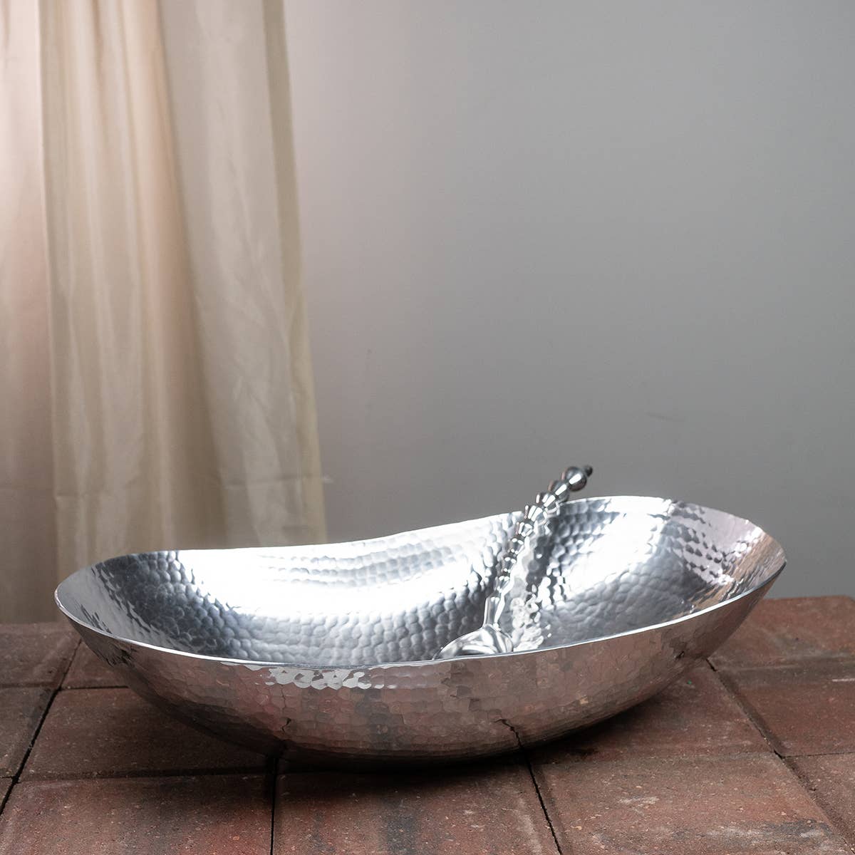 Extra-large hammered silver oval bowl with a matching serving spoon on a brick surface.