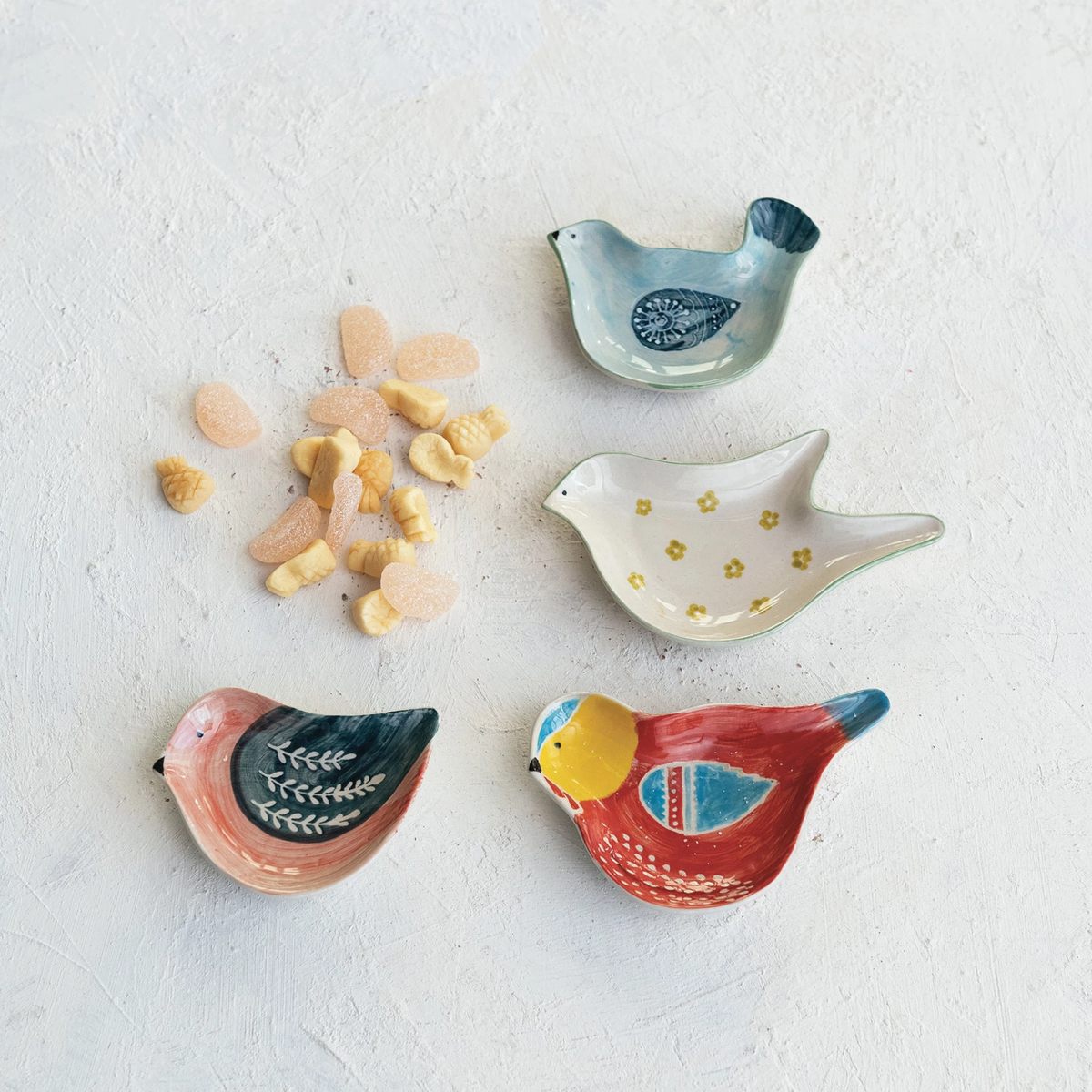 Four hand-painted stoneware bird-shaped dishes in various colors and patterns with small candies.
