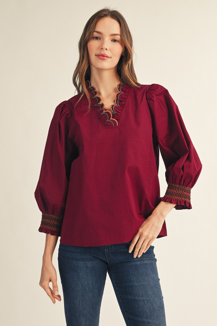 Woman wearing a burgundy top with an embroidered scallop V-neckline and 3/4 puff sleeves.