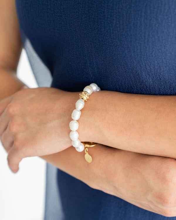 A person wearing a white pearl bracelet with a 24K gold toggle clasp and a small gold charm.