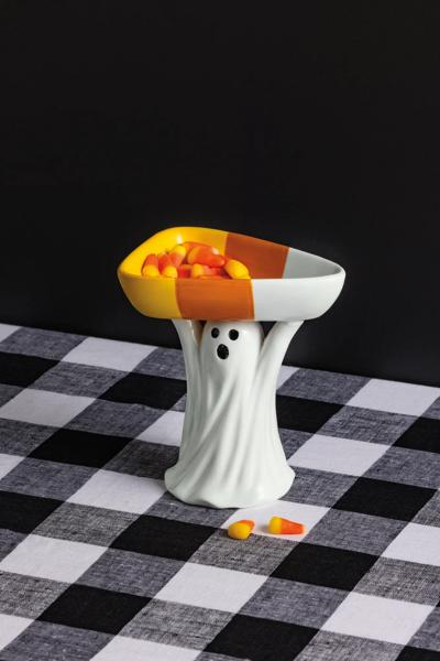 Ceramic ghost figurine holding a candy corn shaped dish filled with candy corn on a checkered cloth.