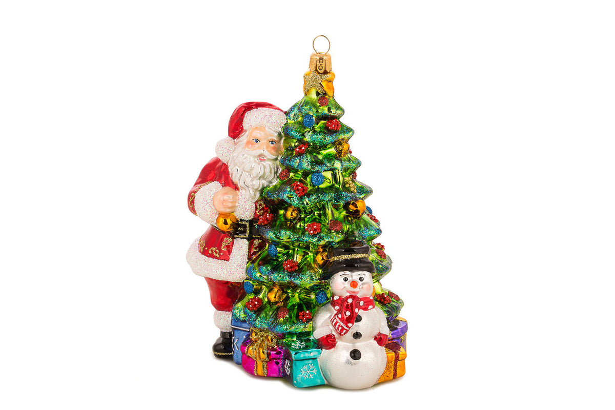 Handcrafted glass ornament of Santa Claus and a snowman beside a decorated Christmas tree.