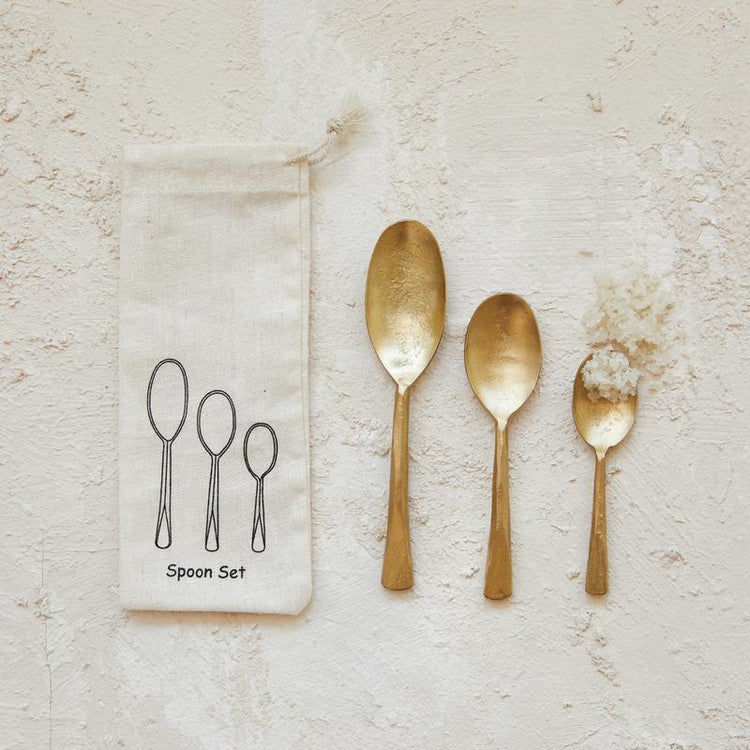 Set of three hand-forged gold-colored iron spoons in varying sizes next to a canvas storage bag.
