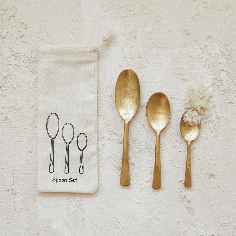 Set of three hand-forged gold-colored iron spoons in varying sizes next to a canvas storage bag.