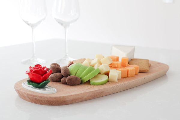 Wooden serving board with a red rose ceramic accent, topped with cheese, fruit, and nuts.