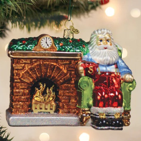 Glass Santa At Hearth Ornament featuring Santa in a green chair next to a brick fireplace.