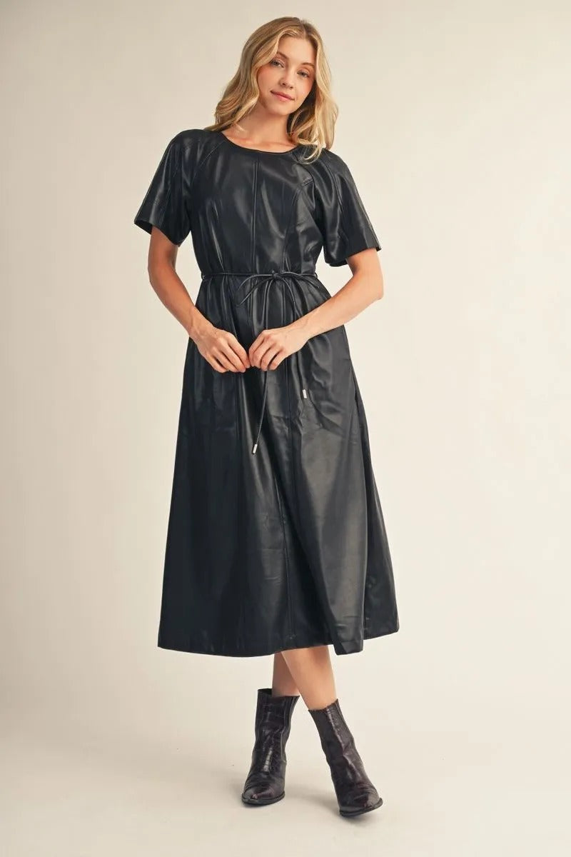 Woman wearing the Allison black short sleeve faux leather midi dress with a tie waist and boots.
