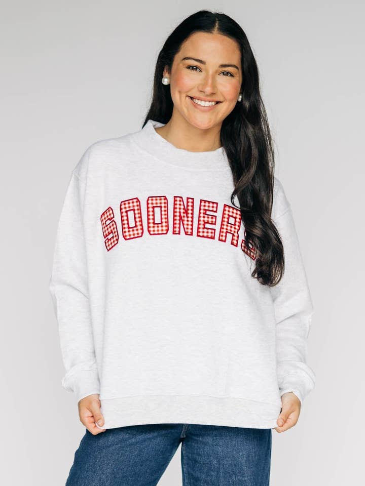 Woman wearing a grey mockneck sweatshirt with 'SOONERS' in red gingham appliqué across the chest.