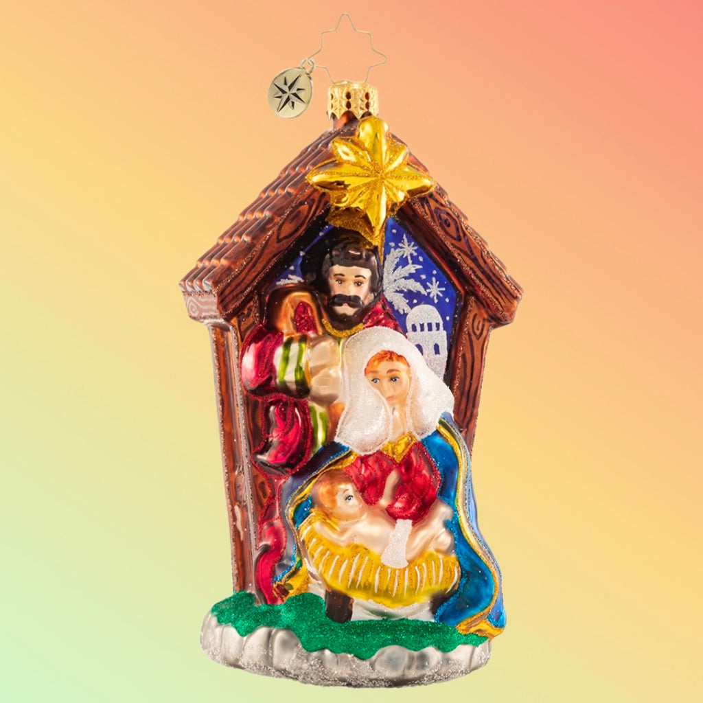 Glass nativity scene ornament featuring Mary, Joseph, and baby Jesus in a brown manger with a star.