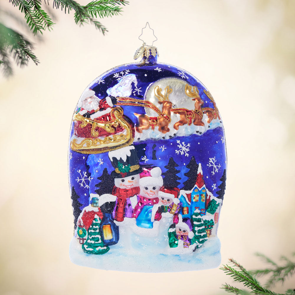 Glass Christmas ornament depicting a snowy village scene with Santa's sleigh and reindeer above.