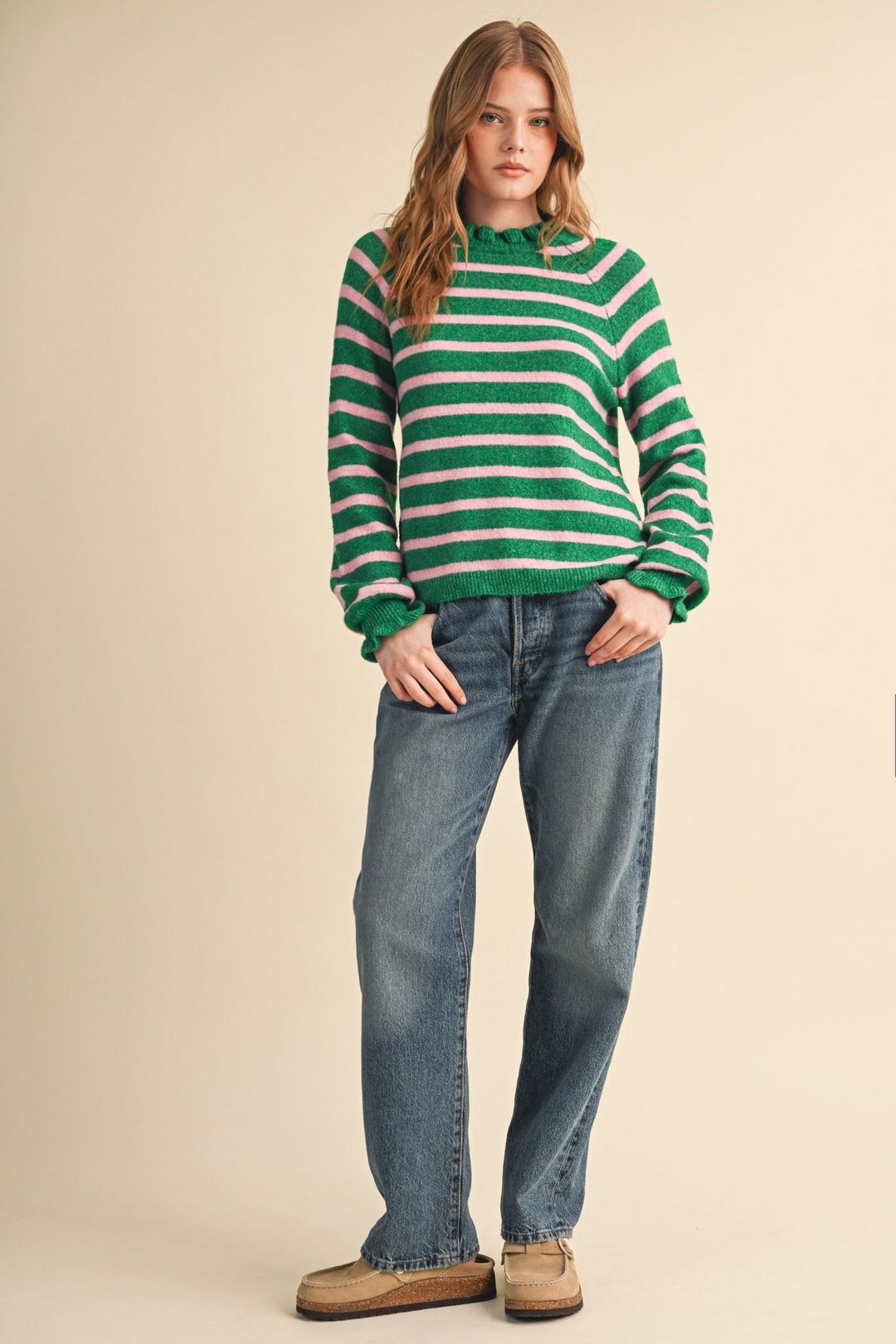 Woman wearing a green and pink striped sweater with ruffled neckline and cuffs with blue jeans.