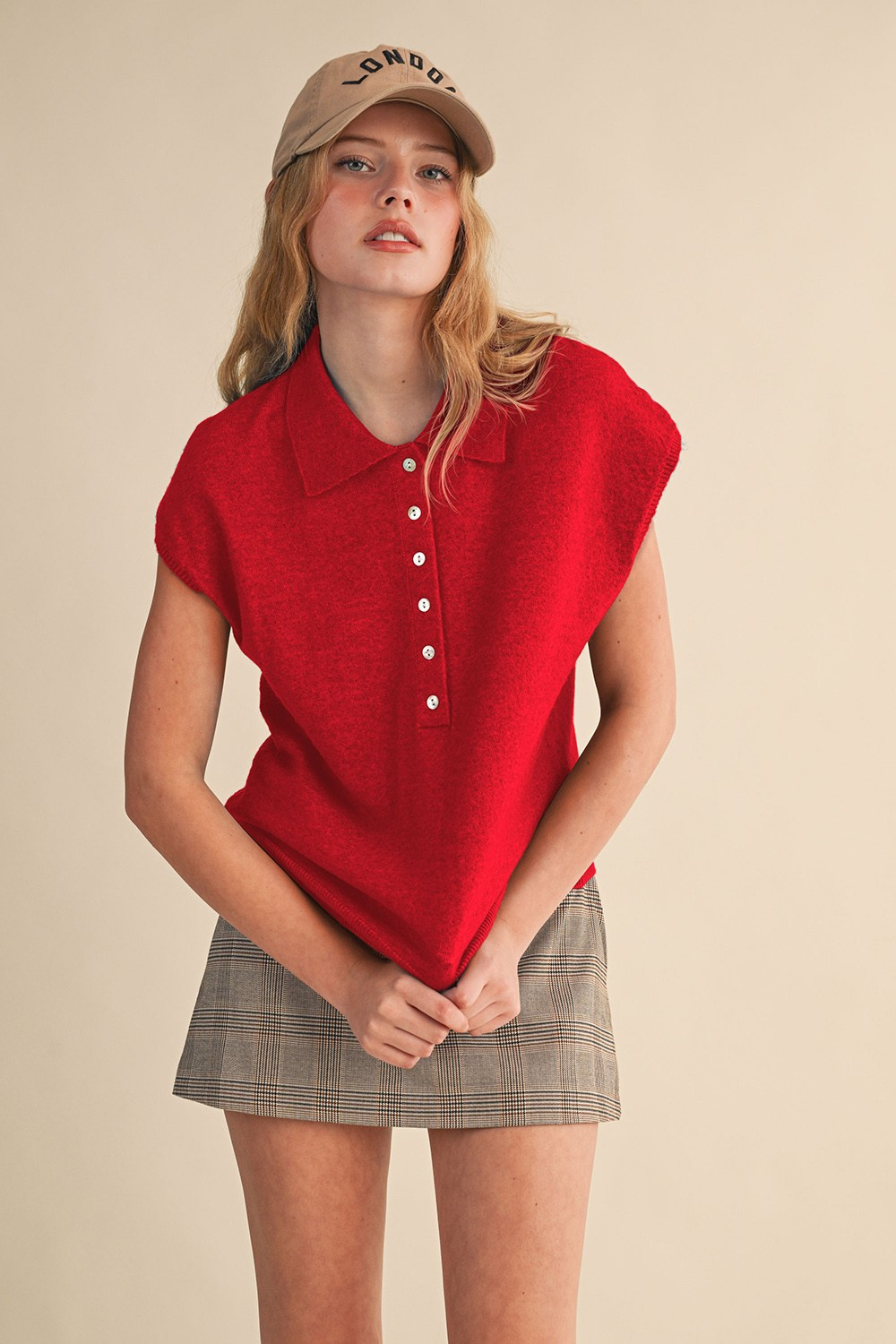Woman wearing a red Jean cap sleeve collared sweater vest with a plaid skirt and tan baseball cap.