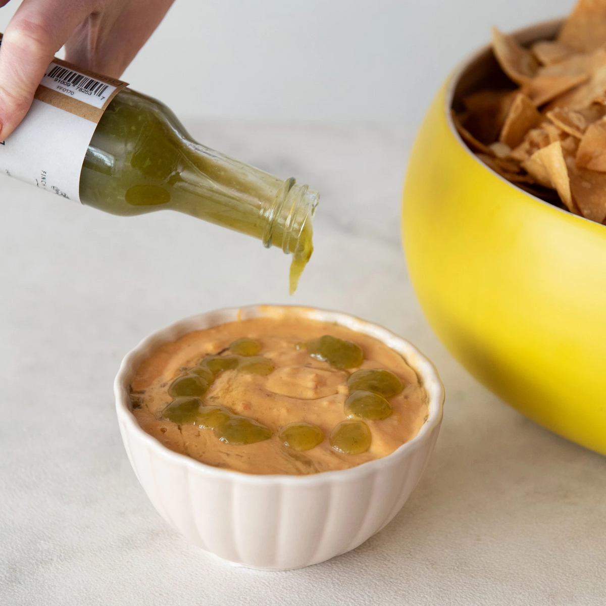 A hand pouring green Verde Hot Sauce from a 5 oz glass bottle into a bowl of creamy orange dip.