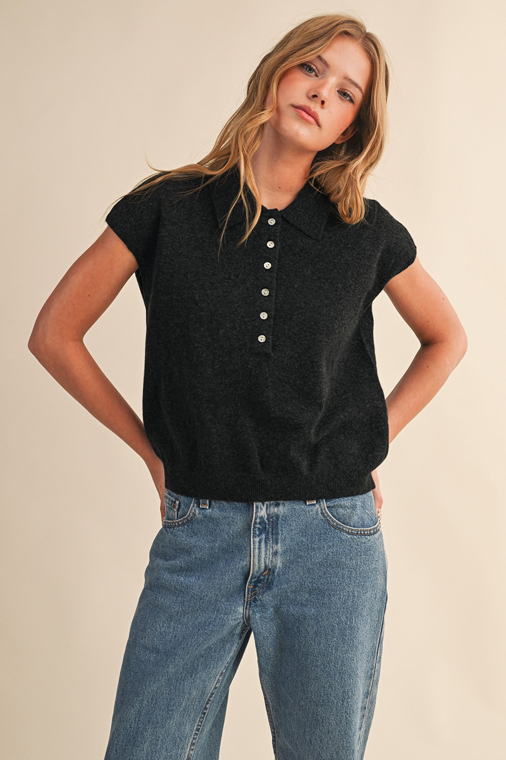 Woman wearing a charcoal grey cap sleeve collared sweater vest with white buttons and blue jeans.