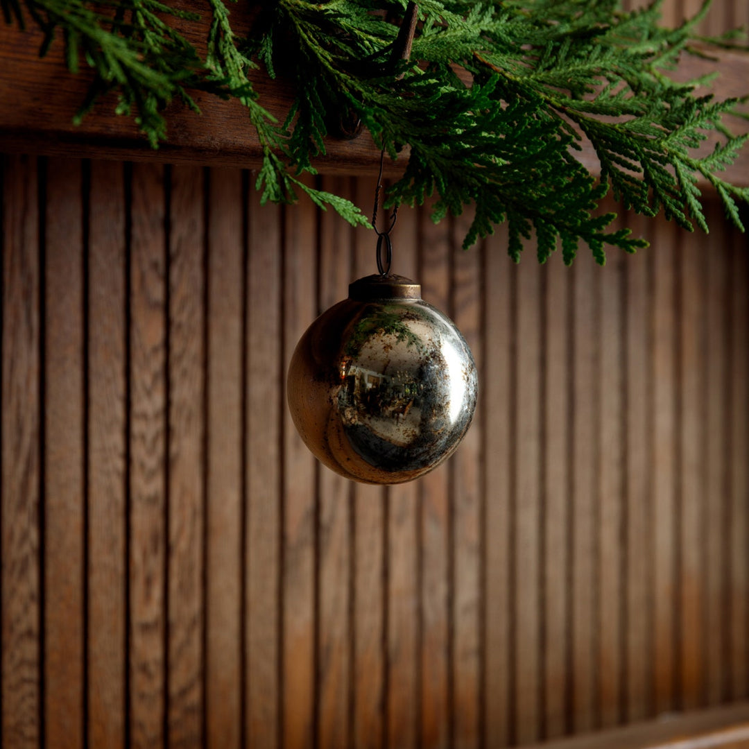 A 3-inch silver mercury glass ball ornament hanging from a green cedar branch against wood paneling.