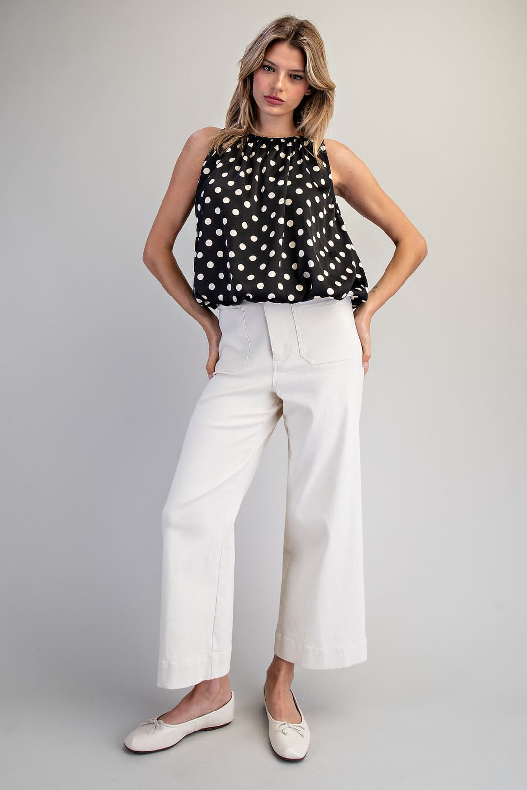 Woman wearing the Monique Polka Dot Sleeveless Blouse in black with white dots and white pants.