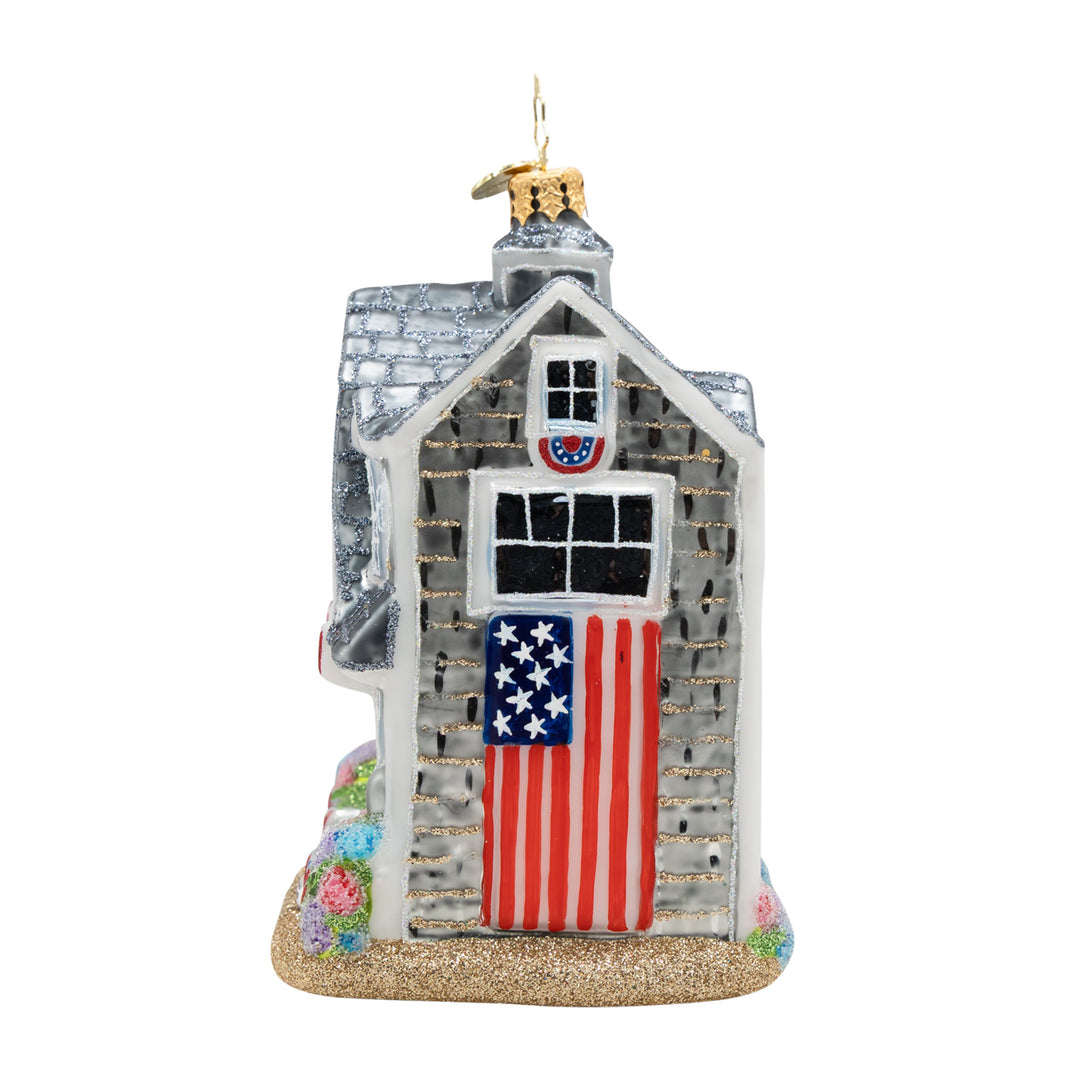 Glass New England beach house ornament with an American flag door and glittery sand base.