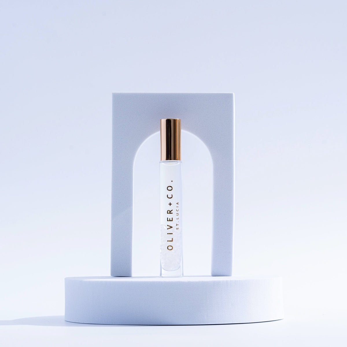 Oliver + Co. Bougie Stick roll-on perfume in a clear glass vial with a gold cap on a white pedestal.
