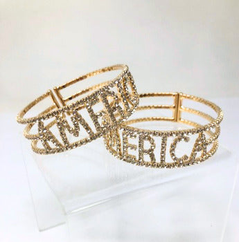Two gold stretch cuff bracelets featuring the word AMERICA in clear rhinestones.