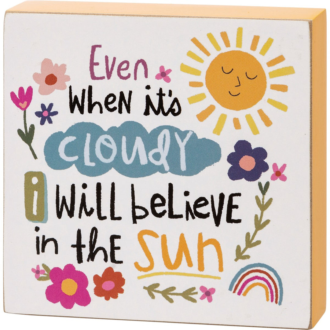 Wooden box sign with "Even When it's Cloudy I Will Believe in the Sun" text, sun, clouds, flowers, and rainbow.