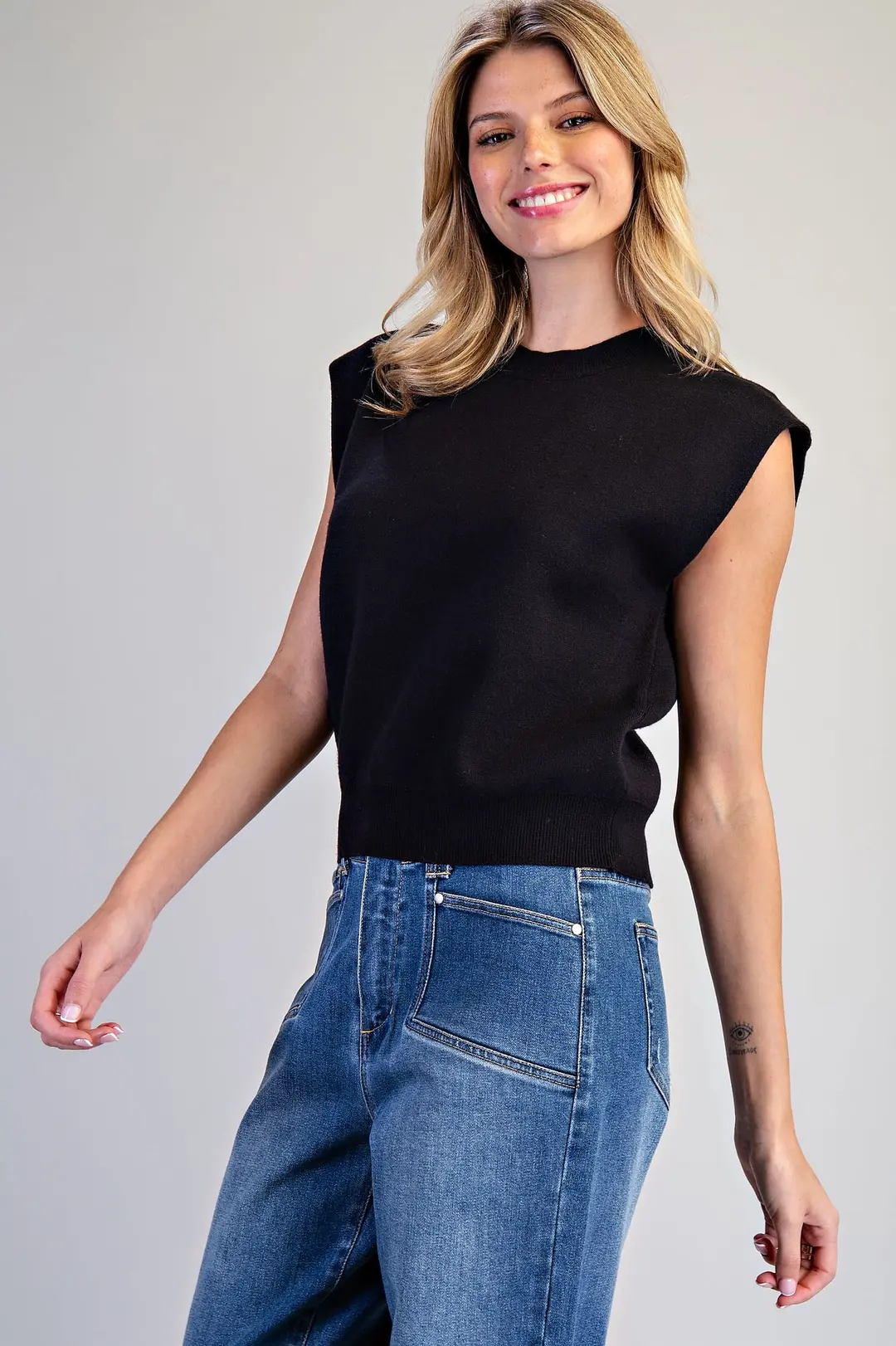 Woman wearing a black sleeveless knit top with extended shoulders and blue jeans.