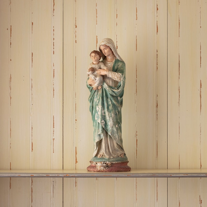 Aged resin Madonna and Child statue with a green robe, standing on a shelf against a wood wall.
