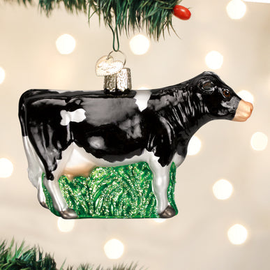 Black and white dairy cow glass ornament standing on green grass, hanging from a Christmas tree.