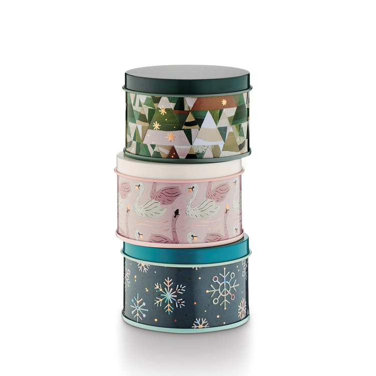 Stack of three round tin gift boxes with festive holiday designs: geometric trees, swans, and snowflakes.