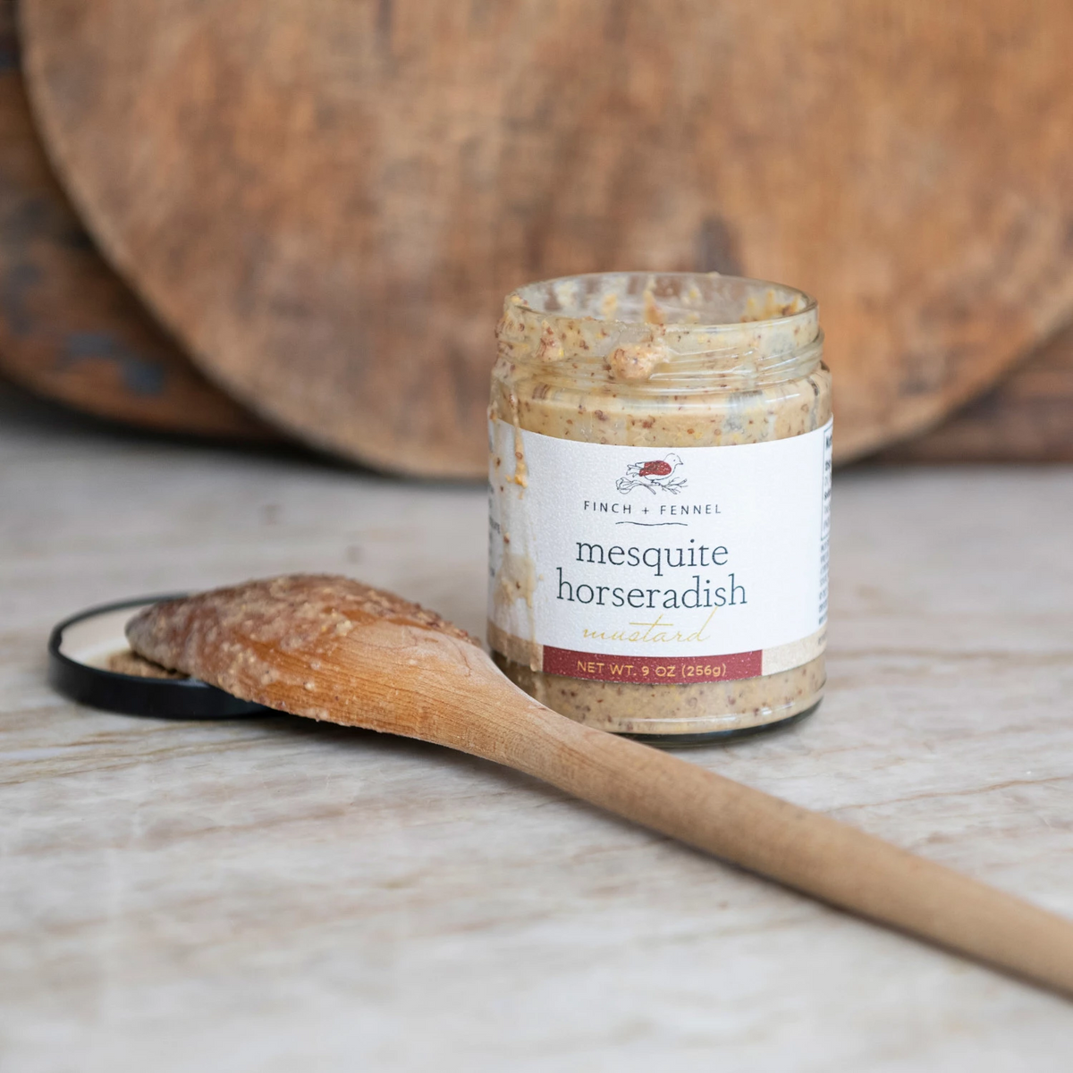 A jar of Finch + Fennel Mesquite Horseradish Mustard next to a wooden spoon with mustard on it.