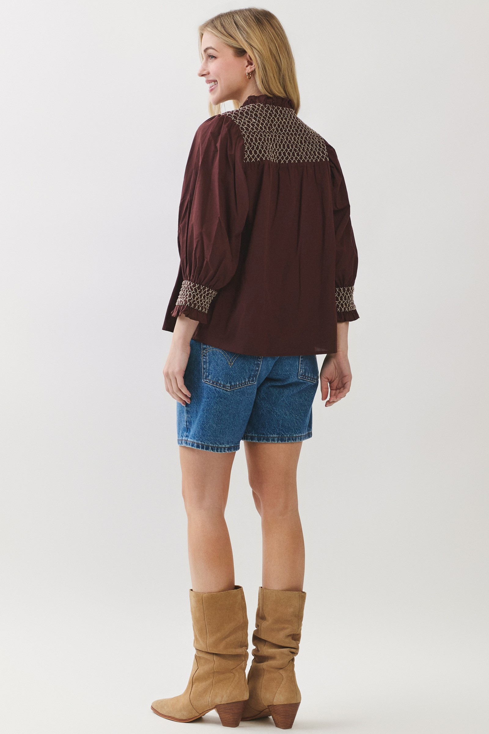 Back view of a woman wearing a maroon Jenny Embroidered Blouse with denim shorts and tan boots.