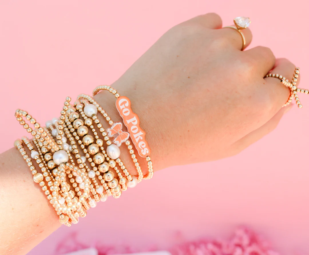 A stack of gold and pearl beaded bracelets on a wrist, featuring an orange 'Go Pokes' charm.