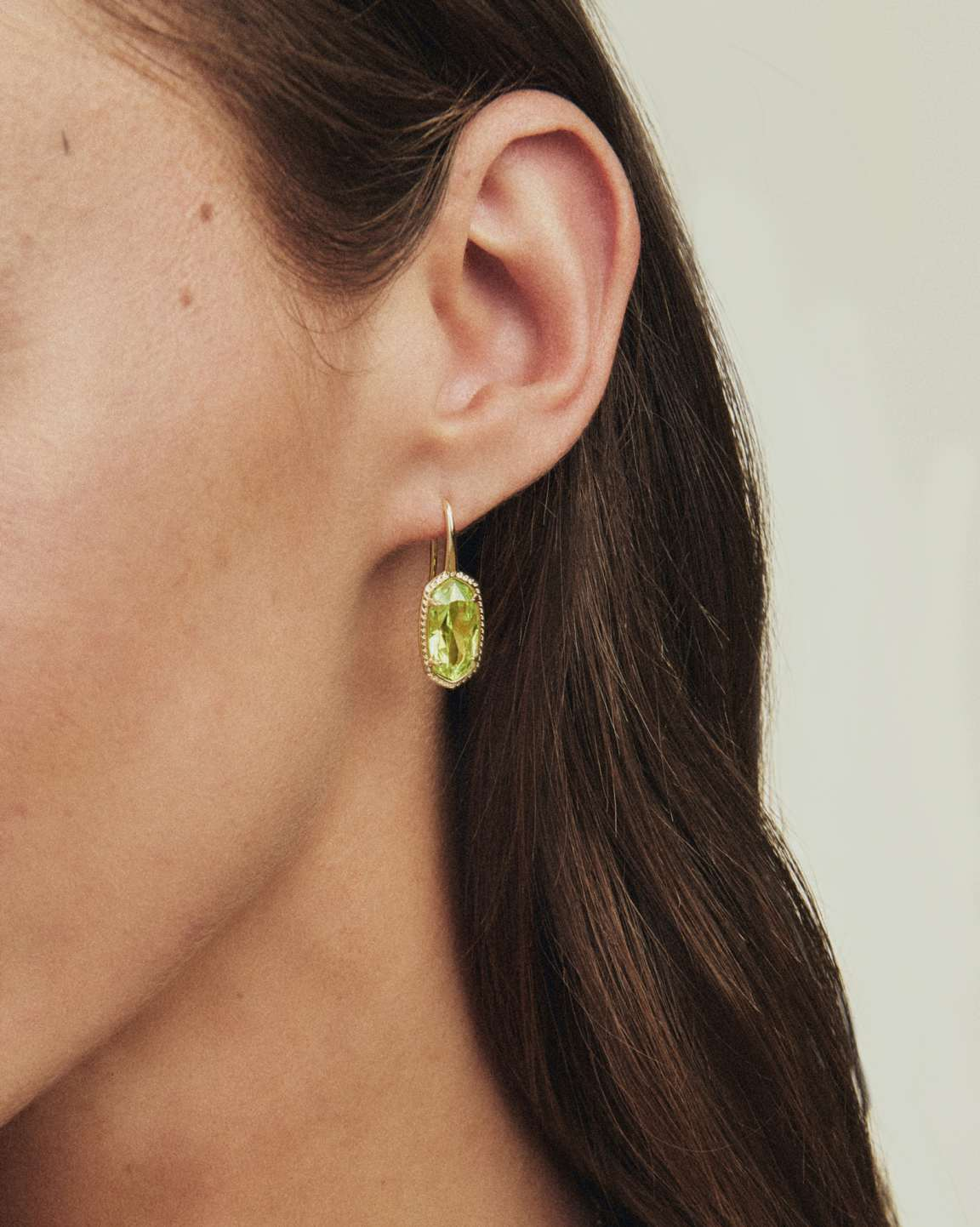 Close-up of a person wearing a Lee Luxe Drop Earring with a faceted green stone and gold setting.