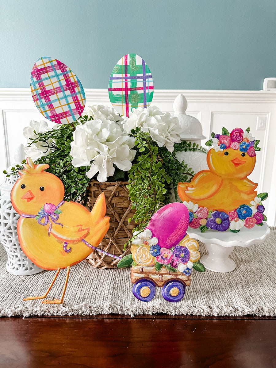 Yellow chick figurine with purple bow, standing next to a flower-filled cart with a pink egg.