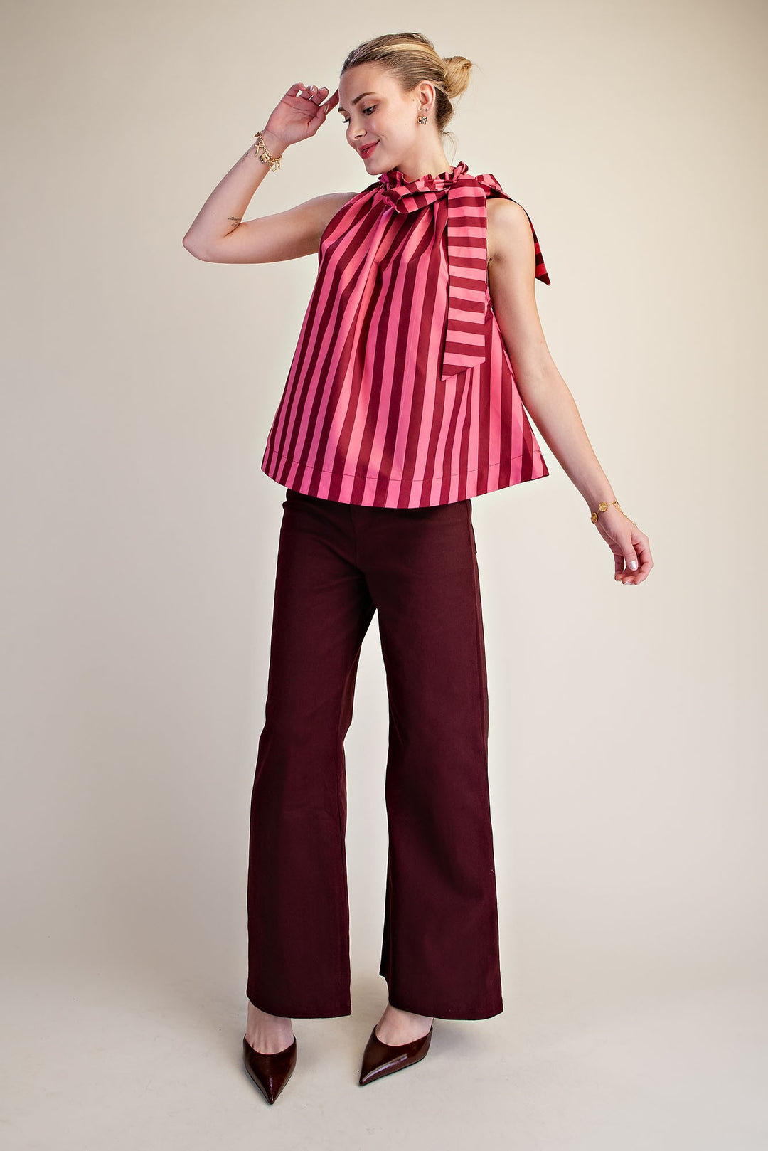 Woman wearing a pink and red vertical stripe sleeveless top with a ruffled tie neck and dark pants.