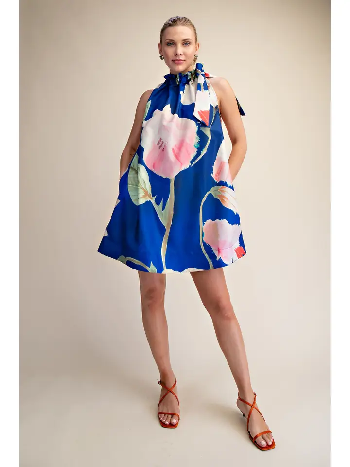 Woman wearing a blue sleeveless mini dress with large pink floral print and a ruffled tie neck.