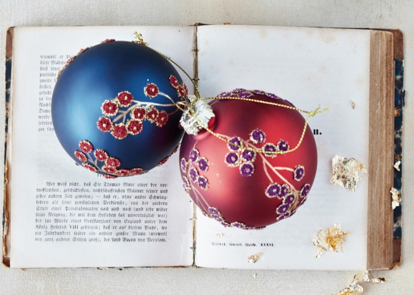Navy and red glass ball ornaments with beaded botanical motifs resting on an open vintage book.