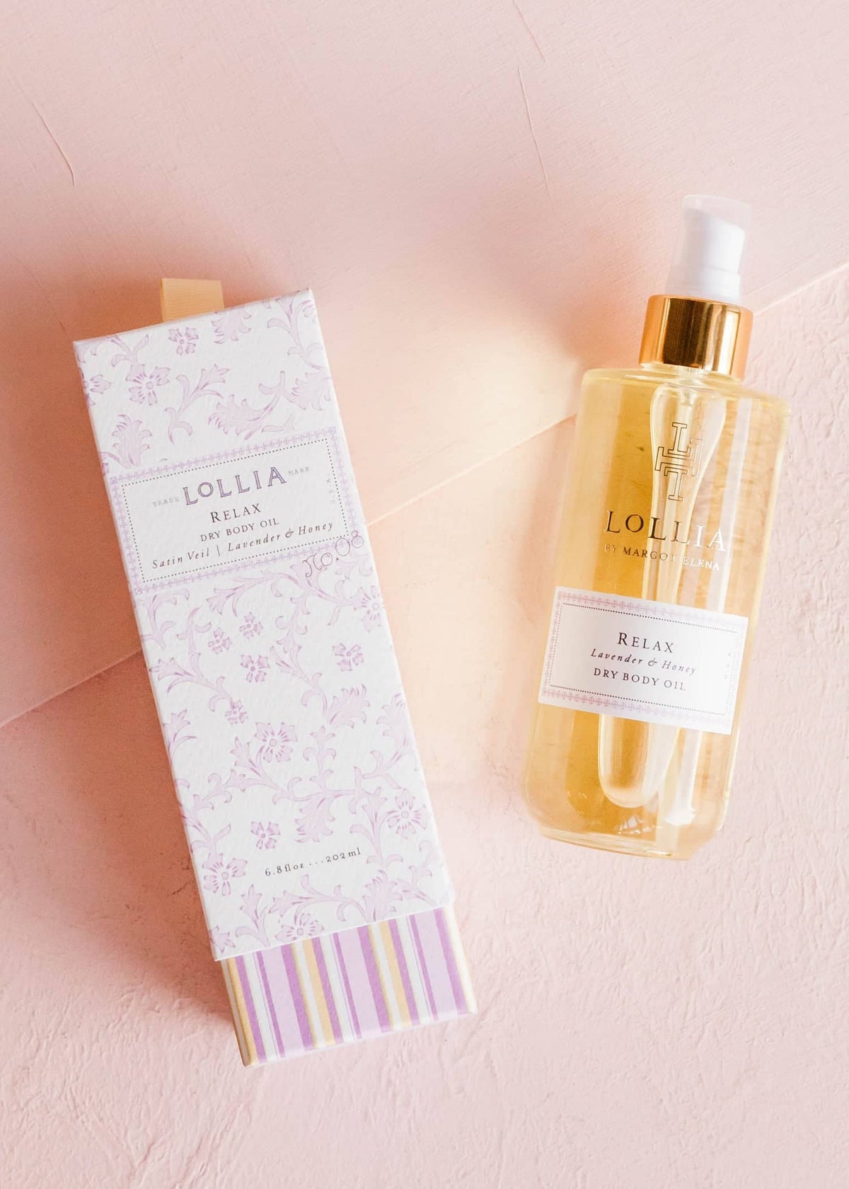 Relax Dry Body Oil in a glass bottle with gold pump next to its floral and striped gift box.