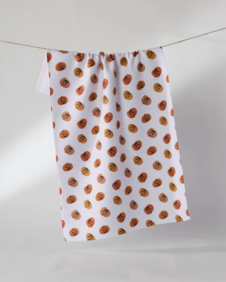 White waffle weave tea towel with an all-over orange jack-o'-lantern pattern hanging on a line.