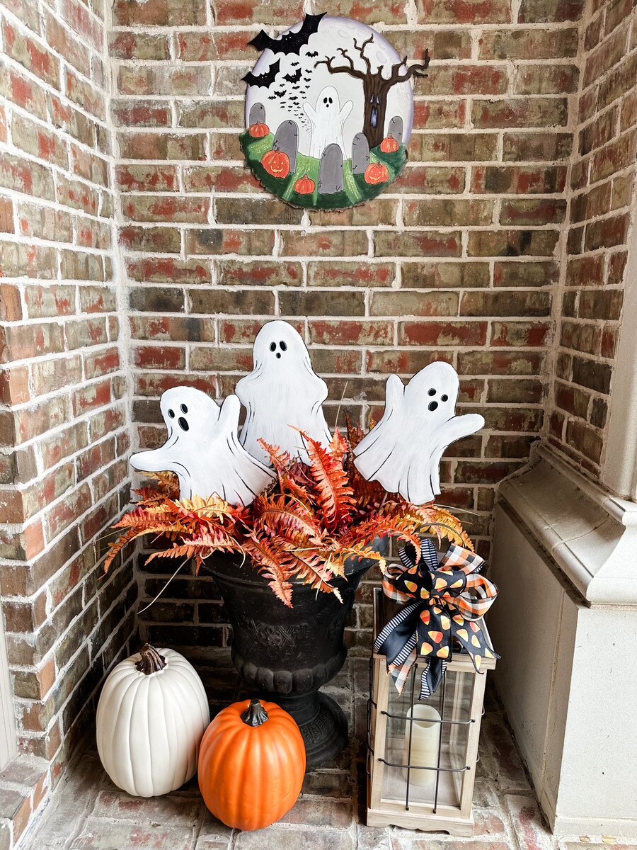 Three white wooden ghost stakes with black outlines and faces in a planter with fall foliage.