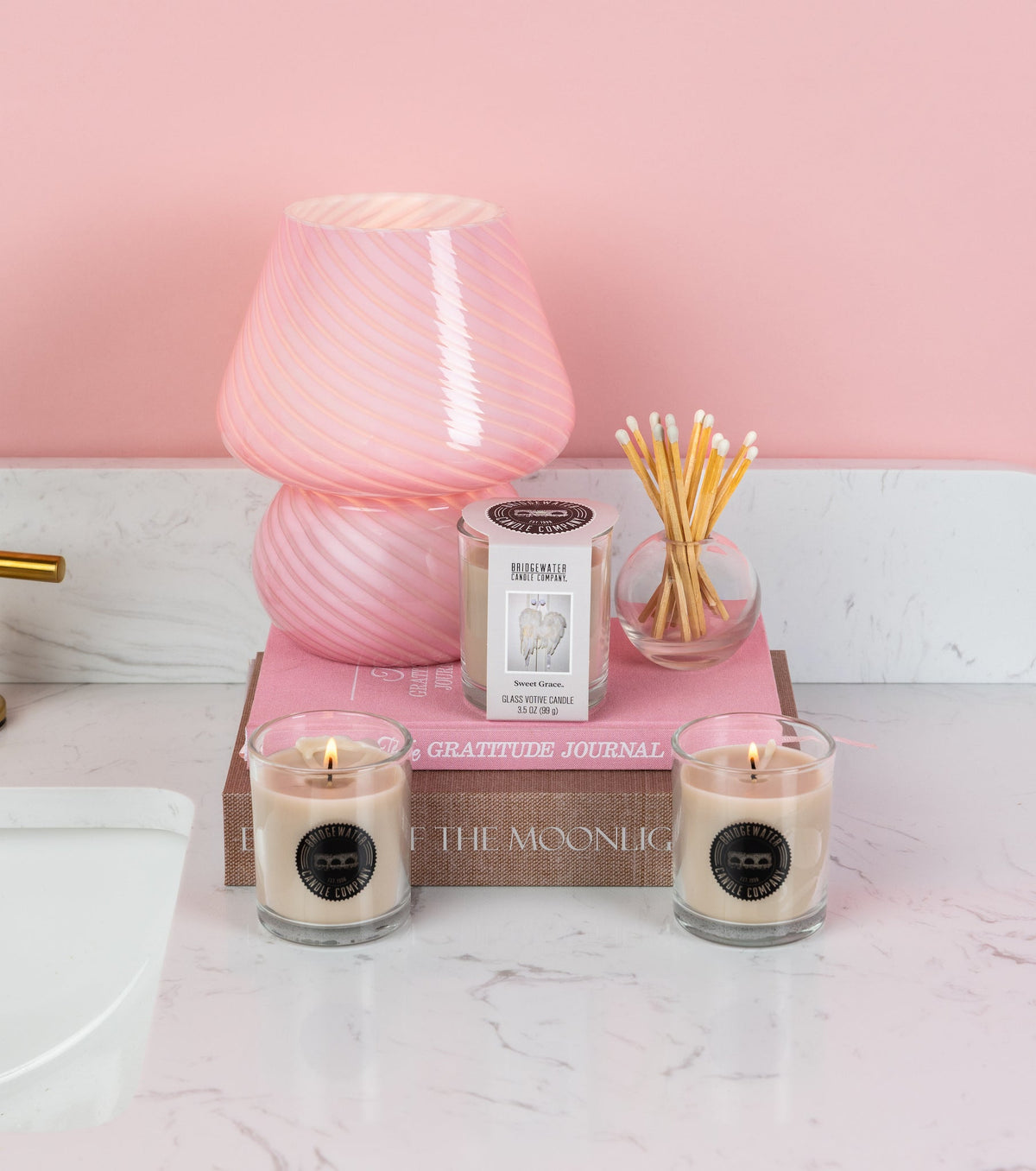 Three Sweet Grace glass votive candles on a pink marble surface with a pink lamp and books.
