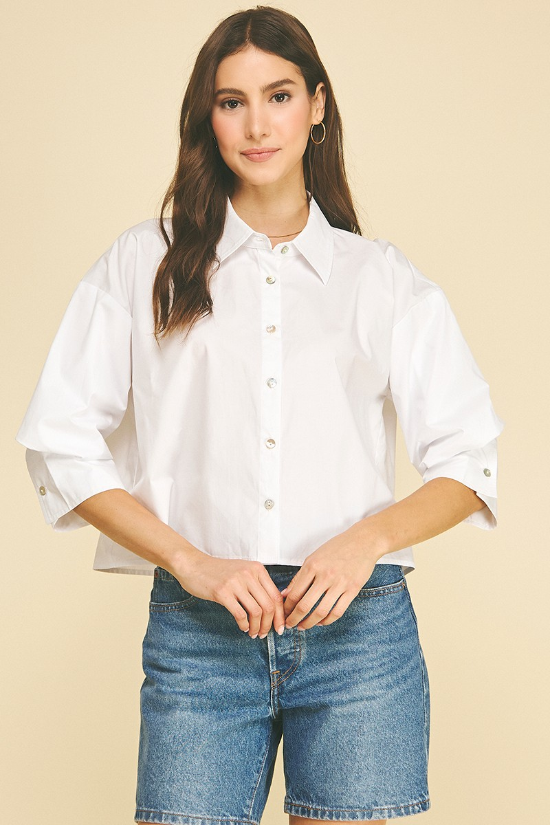 Woman wearing a white cropped button-down shirt with 3/4 tuck detailed sleeves and denim shorts.