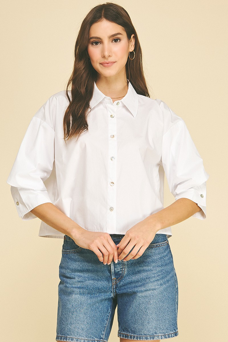 Woman wearing a white cropped button-down shirt with 3/4 tuck detailed sleeves and denim shorts.