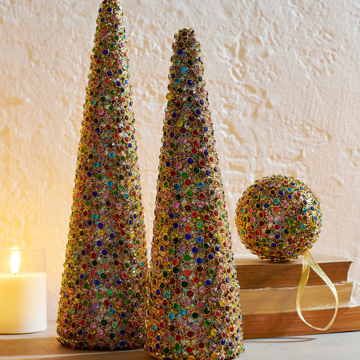 Two multicolor jewel-encrusted cone trees and a matching ornament on a stack of books.