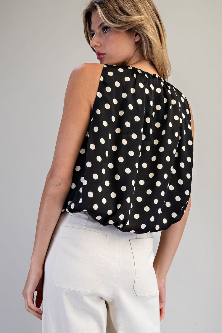 Back view of a woman wearing a black sleeveless blouse with white polka dots and white pants.